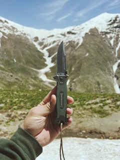 A rugged hand holding a sharp bushcraft knife against a backdrop of forest greenery.