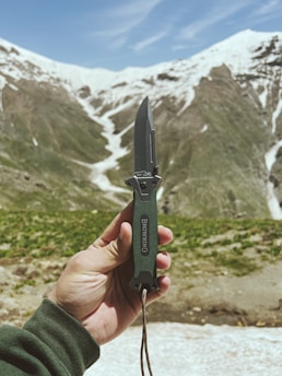 A rugged hand holding a sharp bushcraft knife against a backdrop of forest greenery.