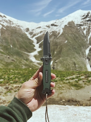 A hand holding a folding knife against a background of snow-capped mountains. The knife has a dark green handle and a metallic blade. The mountains have green patches and snow-covered peaks under a blue sky.