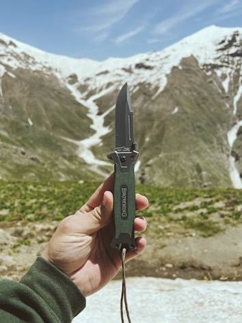A hand holding a folding knife against a background of snow-capped mountains. The knife has a dark green handle and a metallic blade. The mountains have green patches and snow-covered peaks under a blue sky.