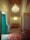 An intricately designed room with an ornate chandelier hanging from a high, decorative ceiling. The room features a mix of traditional and luxurious elements, including a patterned rug, upholstered chairs, and richly detailed white plasterwork on the walls and ceiling. A bright green door and a tapestry with blue geometric patterns add vibrant colors to the calm beige and cream tones of the room.