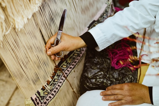 A person wearing a white long-sleeve garment is working on a textile project. They hold a pair of scissors, trimming threads on a loom where a detailed pattern with earthy tones is being created. Vivid pink yarn is visible alongside a black plastic bag.