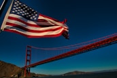 An American flag waves prominently in the foreground with the Golden Gate Bridge extending across the middle of the image. The sky is clear and blue, with a few small birds visible in the distance. The surrounding landscape includes hills and water beneath the bridge.
