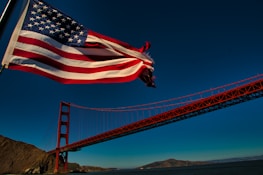 An American flag waves prominently in the foreground with the Golden Gate Bridge extending across the middle of the image. The sky is clear and blue, with a few small birds visible in the distance. The surrounding landscape includes hills and water beneath the bridge.
