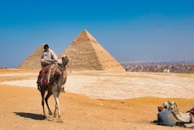 A person is riding a camel in a desert landscape with two large pyramids in the background. The pyramids are located near a city with various buildings visible on the horizon. Another person is seated near a camel on the ground, adjusting or interacting with it.