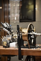 A complex piece of machinery is placed on a wooden table, consisting of various metal components, dials, and copper coils. It appears to be part of a scientific instrument, possibly for measurement or experimentation.