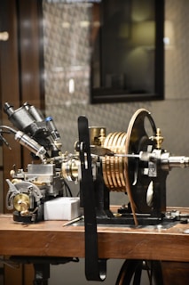A complex piece of machinery is placed on a wooden table, consisting of various metal components, dials, and copper coils. It appears to be part of a scientific instrument, possibly for measurement or experimentation.