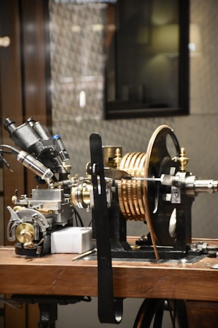 A complex piece of machinery is placed on a wooden table, consisting of various metal components, dials, and copper coils. It appears to be part of a scientific instrument, possibly for measurement or experimentation.
