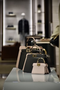 A collection of stylish handbags is displayed on a reflective surface in a well-lit retail environment. There are three handbags of varying sizes, predominantly featuring a black and beige color scheme. In the background, shelves with other products are visible, slightly out of focus, creating a sophisticated and upscale atmosphere.