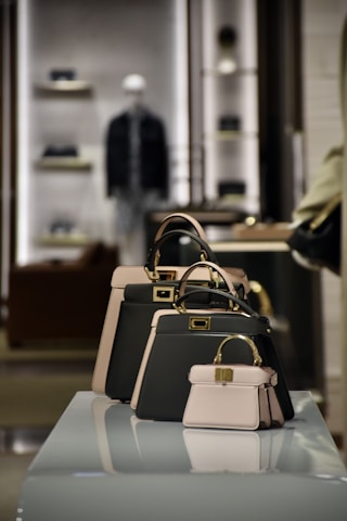 A collection of stylish handbags is displayed on a reflective surface in a well-lit retail environment. There are three handbags of varying sizes, predominantly featuring a black and beige color scheme. In the background, shelves with other products are visible, slightly out of focus, creating a sophisticated and upscale atmosphere.