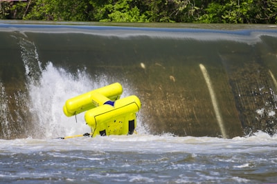 A vibrant yellow raft is flipping over the edge of a small waterfall or weir. Water splashes forcefully around the raft, indicating strong currents. Dense green foliage forms the backdrop, framing the scene with natural greenery.