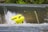 A vibrant yellow raft is flipping over the edge of a small waterfall or weir. Water splashes forcefully around the raft, indicating strong currents. Dense green foliage forms the backdrop, framing the scene with natural greenery.