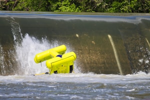 A vibrant yellow raft is flipping over the edge of a small waterfall or weir. Water splashes forcefully around the raft, indicating strong currents. Dense green foliage forms the backdrop, framing the scene with natural greenery.