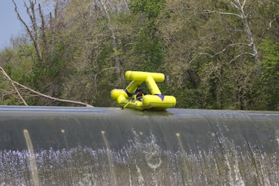 A bright yellow inflatable raft with one person is precariously positioned at the edge of a large waterfall. The surrounding area is dense with lush green trees, and the water flows steadily over the edge.