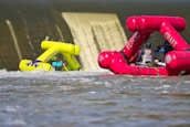 A vibrant yellow inflatable rescue boat appears to be partially submerged in front of a dam's spillway, with water cascading down the structure. Nearby, a pink rescue craft with multiple people on board is heading towards the scene, suggesting an active rescue operation on a flowing river.