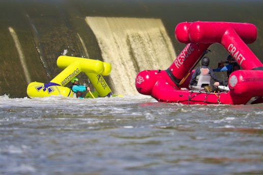 A vibrant yellow inflatable rescue boat appears to be partially submerged in front of a dam's spillway, with water cascading down the structure. Nearby, a pink rescue craft with multiple people on board is heading towards the scene, suggesting an active rescue operation on a flowing river.