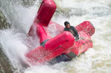 A person wearing a helmet and life jacket maneuvers a red rescue raft through turbulent, white-water rapids. Water splashes forcefully around the raft as it navigates the powerful currents.
