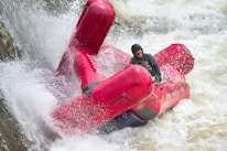 A person wearing a helmet and life jacket maneuvers a red rescue raft through turbulent, white-water rapids. Water splashes forcefully around the raft as it navigates the powerful currents.