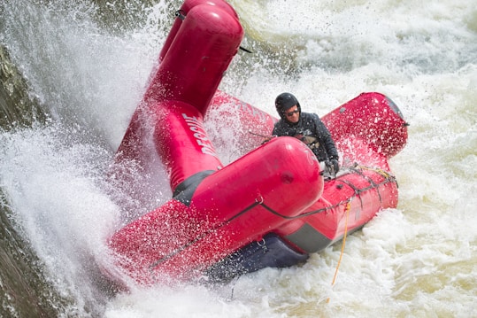 A person wearing a helmet and life jacket maneuvers a red rescue raft through turbulent, white-water rapids. Water splashes forcefully around the raft as it navigates the powerful currents.