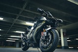 A sleek, modern motorcycle with a black and metallic finish is parked in an urban, dimly lit parking garage. The bike features sporty design elements, including aerodynamic bodywork and prominent disc brakes. Overhead fluorescent lights create a dramatic lighting effect with shadows and highlights on the motorcycle.