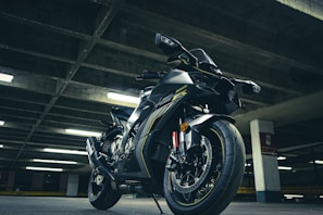 A sleek, modern motorcycle with a black and metallic finish is parked in an urban, dimly lit parking garage. The bike features sporty design elements, including aerodynamic bodywork and prominent disc brakes. Overhead fluorescent lights create a dramatic lighting effect with shadows and highlights on the motorcycle.