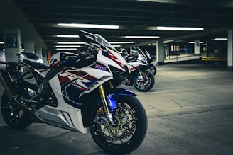 A group of high-performance motorcycles positioned in an underground parking lot, showcasing sleek white, red, and blue racing designs. The concrete surroundings and fluorescent lighting create a dramatic and energetic atmosphere.