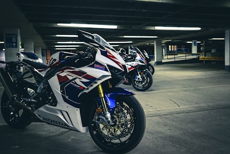 A group of high-performance motorcycles positioned in an underground parking lot, showcasing sleek white, red, and blue racing designs. The concrete surroundings and fluorescent lighting create a dramatic and energetic atmosphere.