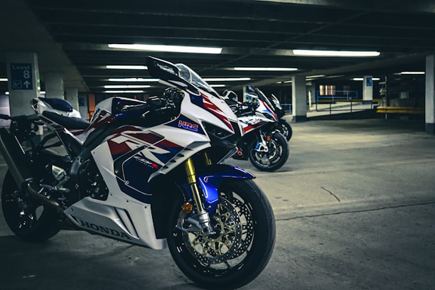 A group of high-performance motorcycles positioned in an underground parking lot, showcasing sleek white, red, and blue racing designs. The concrete surroundings and fluorescent lighting create a dramatic and energetic atmosphere.