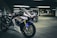 A group of high-performance motorcycles positioned in an underground parking lot, showcasing sleek white, red, and blue racing designs. The concrete surroundings and fluorescent lighting create a dramatic and energetic atmosphere.