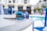 Reflective sunglasses with blue lenses rest on a white table. In the background, there's a swimming pool area with blurred images of chairs and buildings. The reflection in the sunglasses shows palm trees, adding a tropical feel.