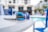 Reflective sunglasses with blue lenses rest on a white table. In the background, there's a swimming pool area with blurred images of chairs and buildings. The reflection in the sunglasses shows palm trees, adding a tropical feel.