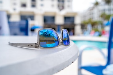 Reflective sunglasses with blue lenses rest on a white table. In the background, there's a swimming pool area with blurred images of chairs and buildings. The reflection in the sunglasses shows palm trees, adding a tropical feel.