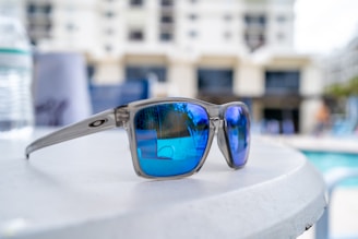 A close-up view of sunglasses with reflective blue lenses resting on a white surface. In the background, there is a blurred image of a modern building and a swimming pool area, creating a relaxed and casual atmosphere.