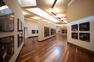 A modern art gallery with wooden flooring and neutral-colored walls, displaying black and white photographs in sleek frames along the walls. The space is well-lit with a combination of natural light from skylights and ceiling-mounted lighting fixtures.