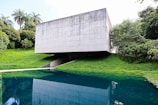 A modern concrete structure with a minimalist design is situated amidst lush greenery. The building is raised above the ground, creating an open space underneath. It overlooks a body of water that reflects the building and the surrounding trees, enhancing the natural setting.