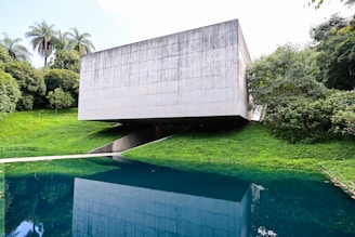 A modern concrete structure with a minimalist design is situated amidst lush greenery. The building is raised above the ground, creating an open space underneath. It overlooks a body of water that reflects the building and the surrounding trees, enhancing the natural setting.