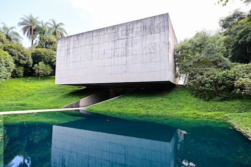 A modern concrete structure with a minimalist design is situated amidst lush greenery. The building is raised above the ground, creating an open space underneath. It overlooks a body of water that reflects the building and the surrounding trees, enhancing the natural setting.
