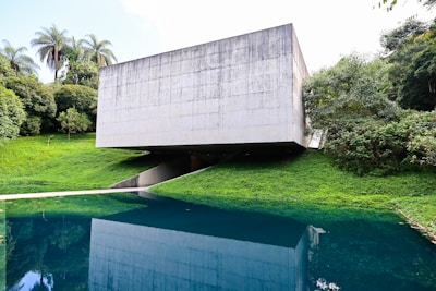 A modern concrete structure with a minimalist design is situated amidst lush greenery. The building is raised above the ground, creating an open space underneath. It overlooks a body of water that reflects the building and the surrounding trees, enhancing the natural setting.