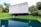 A modern concrete structure with a minimalist design is situated amidst lush greenery. The building is raised above the ground, creating an open space underneath. It overlooks a body of water that reflects the building and the surrounding trees, enhancing the natural setting.