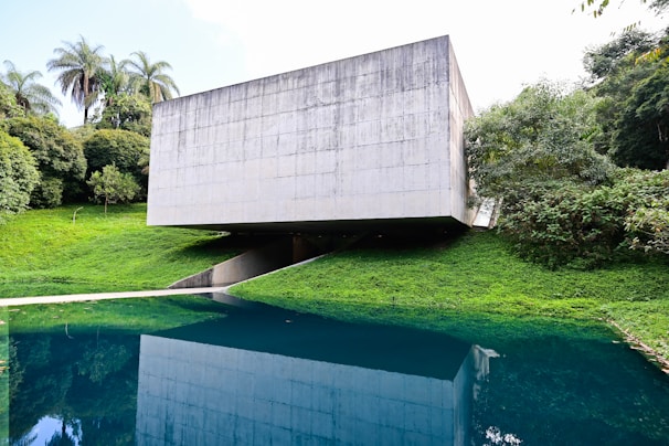 A modern concrete structure with a minimalist design is situated amidst lush greenery. The building is raised above the ground, creating an open space underneath. It overlooks a body of water that reflects the building and the surrounding trees, enhancing the natural setting.