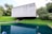 A modern concrete structure with a minimalist design is situated amidst lush greenery. The building is raised above the ground, creating an open space underneath. It overlooks a body of water that reflects the building and the surrounding trees, enhancing the natural setting.