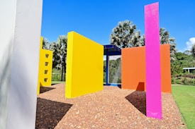 Colorful geometric structures stand in a garden setting. Brightly painted walls in vivid yellow, pink, orange, and blue form a playful outdoor installation. The structures are surrounded by lush greenery, including palm trees under a clear blue sky. The ground is covered in reddish-brown gravel.
