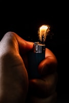 A hand is gripping a lighter with a small, bright flame illuminated against a dark background. The flame casts a warm glow on the fingers, creating a contrast between the light of the flame and the shadowy surrounding. The body of the lighter is metallic and slightly reflective.