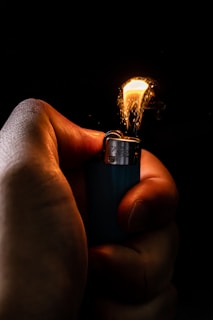 A hand is gripping a lighter with a small, bright flame illuminated against a dark background. The flame casts a warm glow on the fingers, creating a contrast between the light of the flame and the shadowy surrounding. The body of the lighter is metallic and slightly reflective.