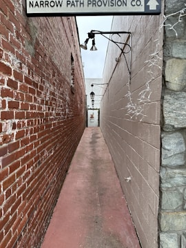 A narrow alleyway formed by two brick and stone walls, leading to a door at the end. Overhead lights are mounted on the walls, and a sign above the entrance displays the name 'Narrow Path Provision Co.' The floor is a faded pinkish-red concrete, and strings of lights are draped along one of the walls.