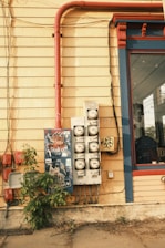 The image depicts an urban scene featuring the side of a building with yellow wooden siding. There are multiple utility meters and electrical boxes mounted on the wall, with some graffiti and stickers. A bright red pipe runs vertically alongside the building, contrasting with the yellow wall. A glass window with a blue and red frame is visible to the right. A small bush grows at the base of the wall.