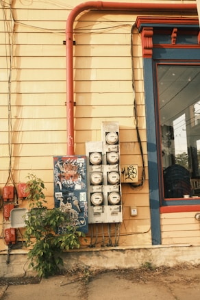 The image depicts an urban scene featuring the side of a building with yellow wooden siding. There are multiple utility meters and electrical boxes mounted on the wall, with some graffiti and stickers. A bright red pipe runs vertically alongside the building, contrasting with the yellow wall. A glass window with a blue and red frame is visible to the right. A small bush grows at the base of the wall.