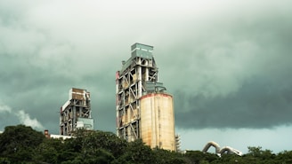 Two large industrial buildings with exposed metal frameworks stand against a backdrop of dark, ominous clouds. Vegetation surrounds the lower part of the structures, suggesting a juxtaposition between industry and nature.