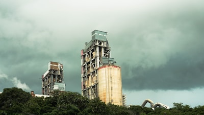 Two large industrial buildings with exposed metal frameworks stand against a backdrop of dark, ominous clouds. Vegetation surrounds the lower part of the structures, suggesting a juxtaposition between industry and nature.