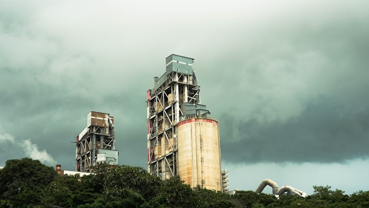 Two large industrial buildings with exposed metal frameworks stand against a backdrop of dark, ominous clouds. Vegetation surrounds the lower part of the structures, suggesting a juxtaposition between industry and nature.